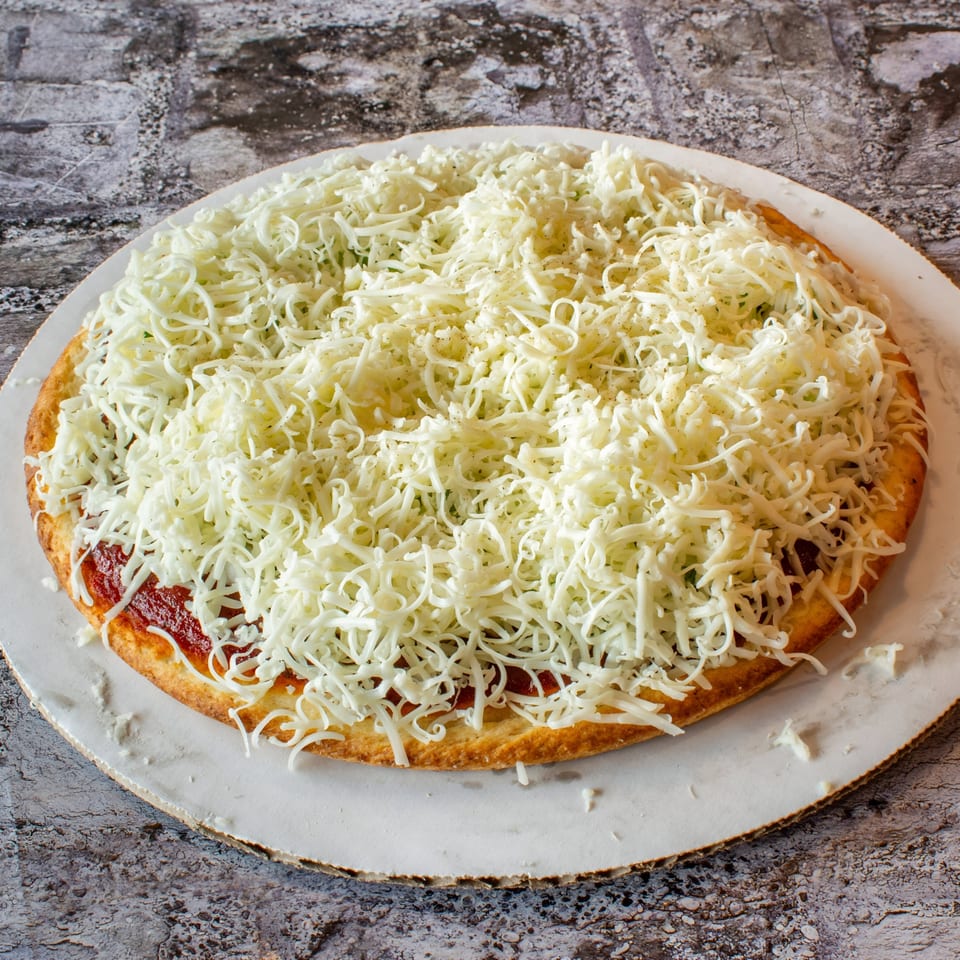 Uncooked Stavro's Pizza House IV pizza with a thick layer of shredded mozzarella cheese.